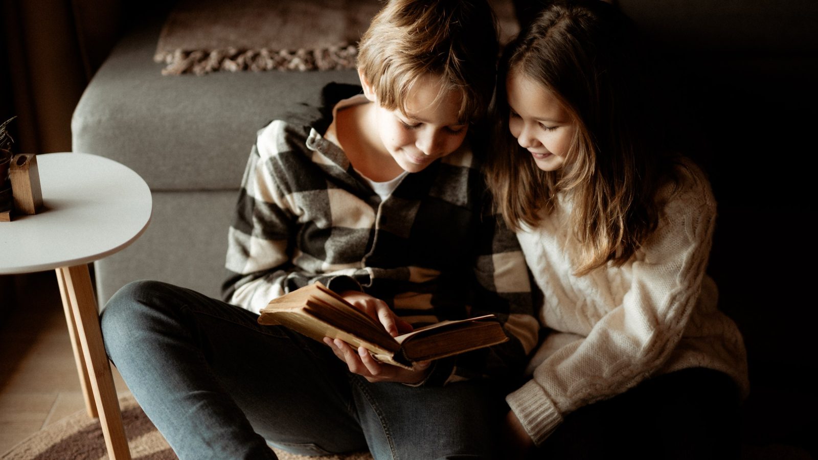 Imagen editorial para «Leer en familia sigue gustando; sostener el momento se vuelve más difícil»: Two children sit closely on a soft surface, engrossed in a book. The room is warmly lit, creating a cozy atmosphere as they share this quiet